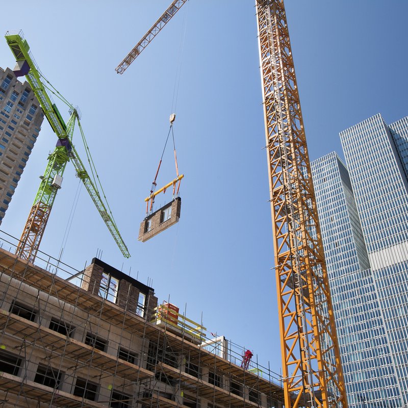 Construction site in urban setting with two tall cranes (green and orange) lifting a building component, showing a partially completed structure with scaffolding in the foreground and several high-rise buildings